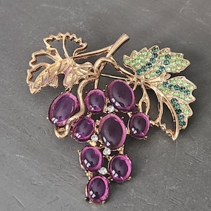 May include: A gold-toned brooch in the shape of a grape cluster. The brooch features oval, purple-colored stones representing the grapes, with green and clear accents on the leaves. The brooch is a decorative accessory.