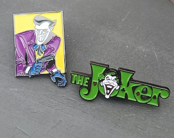 May include: Two enamel pins featuring The Joker. One pin shows the Joker in a purple suit, holding a gun, set against a yellow background. The other pin spells "The Joker" in green with the Joker's face.