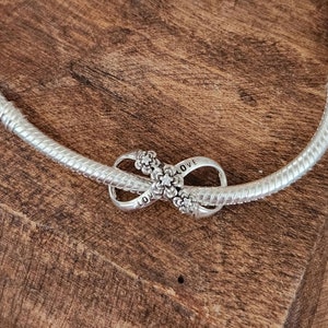 May include: A silver-colored necklace with a snake chain design. The necklace features an infinity charm with the word "LOVE" inscribed on it, adorned with floral accents. The jewelry is displayed on a wooden surface.