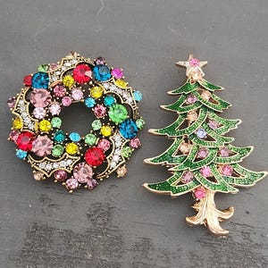 May include: Two decorative brooches: a wreath and a Christmas tree. The wreath brooch is gold-toned, adorned with colorful gemstones in shades of red, blue, pink, and yellow. The Christmas tree brooch is green with pink and clear gemstones, topped with a pink star.