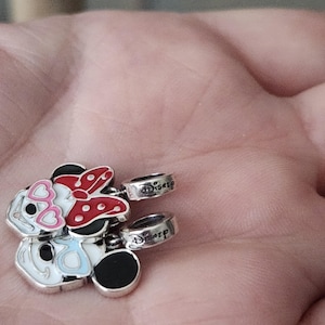 May include: Silver-toned Disney charms featuring Minnie and Mickey Mouse designs. Minnie has a red bow and Mickey has black ears. The charms have the word "Disney" engraved on the rings.