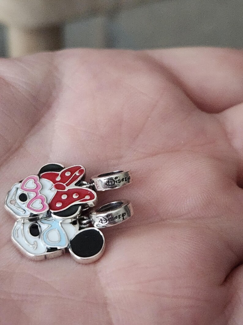 May include: Silver-toned Disney charms featuring Minnie and Mickey Mouse designs. Minnie has a red bow and Mickey has black ears. The charms have the word "Disney" engraved on the rings.