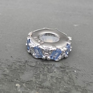 May include: A silver-toned charm bead featuring a circular design with alternating light blue baguette-cut stones and clear round stones. The metal surface is engraved with the text S925, indicating the material composition of the jewelry piece.