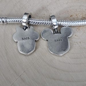 May include: Two silver charms with Mickey Mouse and Minnie Mouse silhouettes, hanging from a silver bracelet. Each charm has the text "S925" stamped on the back. The charms are on a light wood surface.