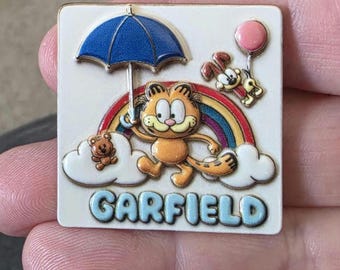 May include: A square enamel pin featuring the orange cartoon cat Garfield holding a blue umbrella under a rainbow. The design includes a small brown teddy bear on a white cloud, a pink balloon, and the name GARFIELD in light blue block letters.