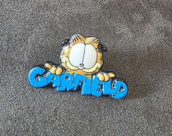 May include: A metal enamel pin featuring the cartoon character Garfield, depicted with orange fur and large white eyes, resting hands on a bright blue banner that displays the name GARFIELD in bold, raised, capital blue lettering.