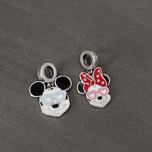 May include: Two silver charms featuring Mickey and Minnie Mouse. Mickey has black ears, blue sunglasses, and a white face. Minnie has black ears, a red and white polka dot bow, and pink heart-shaped sunglasses. Both charms have a silver loop.
