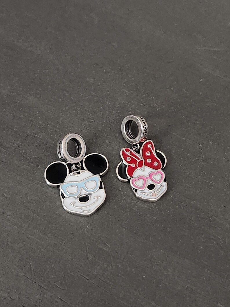 May include: Two silver charms featuring Mickey and Minnie Mouse. Mickey has black ears, blue sunglasses, and a white face. Minnie has black ears, a red and white polka dot bow, and pink heart-shaped sunglasses. Both charms have a silver loop.