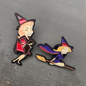 May include: Two enamel pins featuring cartoon witches. One pin shows a witch in a black dress and red cape, holding a red bag. The other pin depicts a witch flying on a broom, wearing a purple cape and dress.
