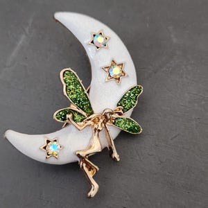 May include: A gold-toned fairy brooch with green glitter wings and a white crescent moon. The moon has iridescent stars and a textured surface. The fairy is perched on the moon.