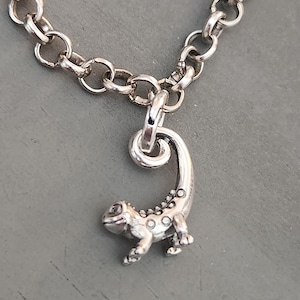 May include: A silver-toned charm necklace featuring a lizard pendant. The lizard has a detailed design with small circular accents along its back and tail. The chain is made of large, rounded links.