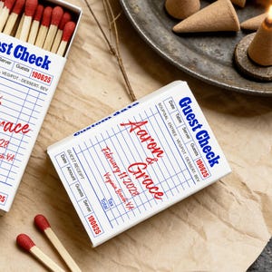 May include: Two white matchboxes featuring a blue and red guest check design. The text reads Guest Check, Aaron & Grace, February 11 2028, and Virginia Beach, VA. One box is open, revealing wooden matches with red tips. The other box is closed.