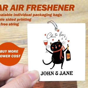 Op de afbeelding: Een witte autoluchtverfrisser met een illustratie van een zwarte kat die een glas wijn en een fles wijn vasthoudt. De tekst "Cheers to love!" en "John & Jane" staat op de luchtverfrisser gedrukt.