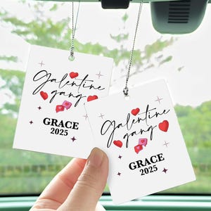 May include: Two white car air fresheners with the text "Galentine gang" in cursive, red hearts, and the name "GRACE" with the year "2025". The air fresheners are suspended by a silver string.