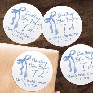 May include: Three round blue and white bridal shower favour stickers with a blue ribbon and the text "Something Blue Before "I do" Maria's bridal shower 11.17.2025"