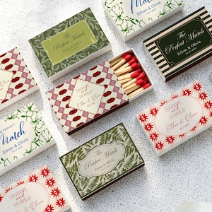 May include: A collection of rectangular matchboxes featuring various decorative patterns, including red lip motifs, green botanical prints, and striped designs. Each box displays the text "The Perfect Match" or "To Have and To Hold" with "Ethan & Olivia" and "08.27.2026".