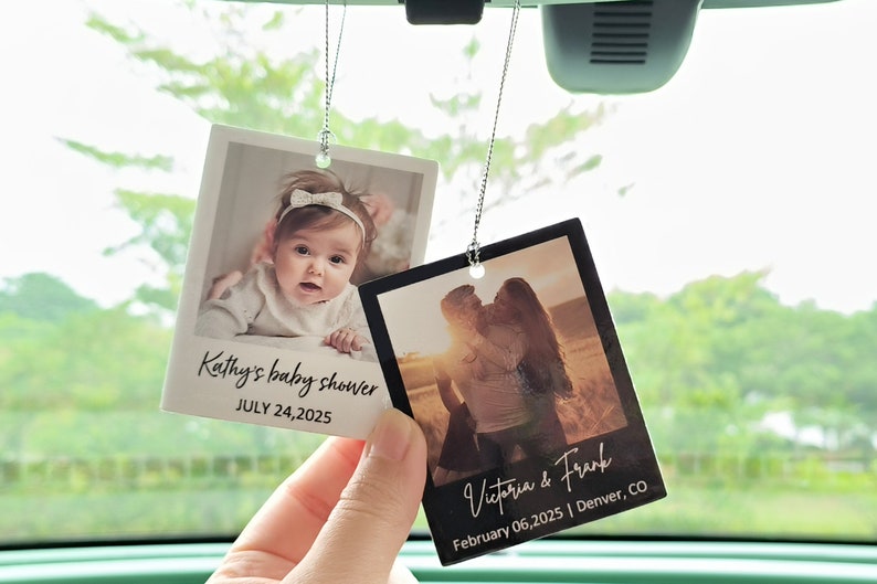 May include: Two photo cards hanging from a rearview mirror in a car. The card on the left features a baby girl and the text "Kathy's baby shower JULY 24, 2025". The card on the right features a couple and the text "Victoria & Frank February 06, 2025 | Denver, CO".