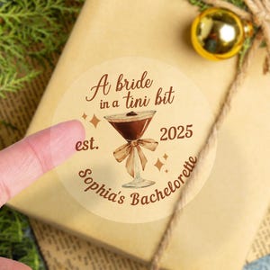 May include: A round sticker with a brown and white cocktail glass illustration. The text reads "A bride in a tini bit est. 2025 Sophia's Bachelorette".