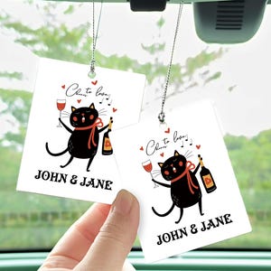 Op de afbeelding: Twee witte autoluchtverfrissers met zwarte katten die wijnglazen en flessen vasthouden. De tekst op de luchtverfrissers luidt "Cheers to love!" en "John & Jane".