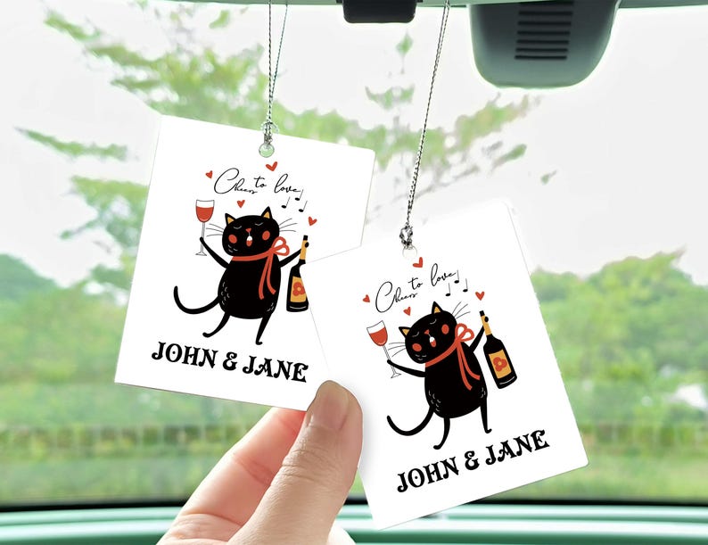 Op de afbeelding: Twee witte autoluchtverfrissers met zwarte katten die wijnglazen en flessen vasthouden. De tekst op de luchtverfrissers luidt "Cheers to love!" en "John & Jane".