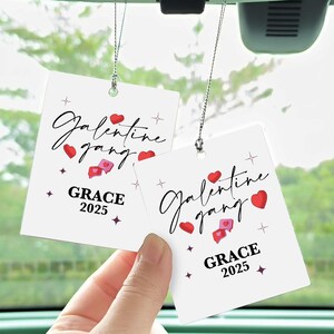 May include: Two white car air fresheners with the text "Galentine gang" in cursive, red hearts, and the name "GRACE" with the year "2025". The air fresheners are suspended by silver string.