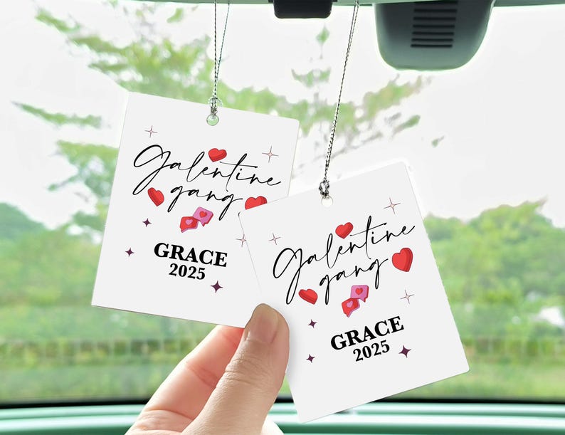 May include: Two white car air fresheners with the text "Galentine gang" in cursive, red hearts, and the name "GRACE" with the year "2025". The air fresheners are suspended by silver string.
