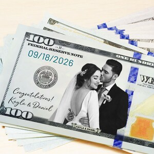 Puede incluir: Una pila de billetes personalizados de 100 d&oacute;lares con tem&aacute;tica de boda. Los billetes muestran una foto de una pareja, la fecha 18/09/2026 y los nombres Kayla & Daniel. Tambi&eacute;n es visible el texto "&iexcl;Felicidades!"