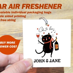 Op de afbeelding: Een witte autoluchtverfrisser met een illustratie van een zwarte kat die een glas wijn en een fles wijn vasthoudt. De tekst "Cheers to love!" en "John & Jane" staat op de luchtverfrisser gedrukt.