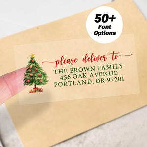May include: A clear sticker featuring a watercolour Christmas tree and the text "please deliver to" followed by an address. A white circle with the text "50+ Font Options" is in the upper right corner. The sticker is on a tan envelope.