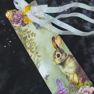 May include: A decorative bookmark featuring a detailed illustration of a rabbit, purple flowers, and butterflies. The bookmark has a white ribbon and a decorative flower and gold leaf accent at the top. The background is a soft green.