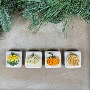Könnte beinhalten: Vier Keramik-Kürbisfliesen in verschiedenen Farben und Designs. Die Fliesen sind quadratisch und haben einen weißen Hintergrund. Die Kürbisse sind in Grün-, Gelb-, Orange- und Brauntönen bemalt.