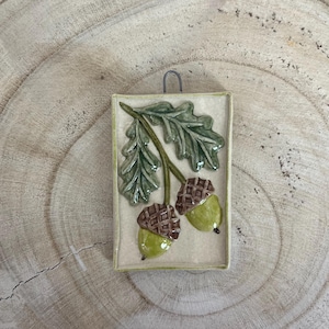 Peut inclure: Un carreau en céramique avec un motif en relief de deux glands et une feuille de chêne. Les glands sont brun et vert, et la feuille est verte. Le carreau est posé sur une surface en bois brun clair.