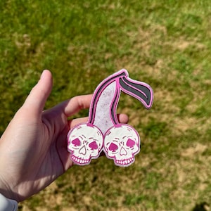 May include: A cherry-shaped patch featuring two white skulls with pink heart-shaped noses and pink teeth. The stems are light purple with a glittery texture and black outlines. The design is flat and has a distinct border around the entire shape.