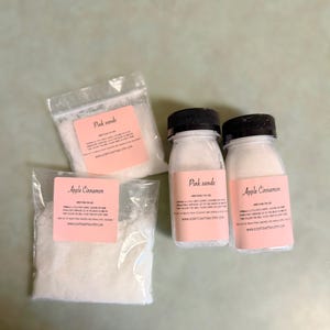 May include: Three small bottles of bath salts, two labelled "Pink Sands" and one labelled "Apple Cinnamon". The bottles are clear and contain white bath salts. The labels are pink with white text.