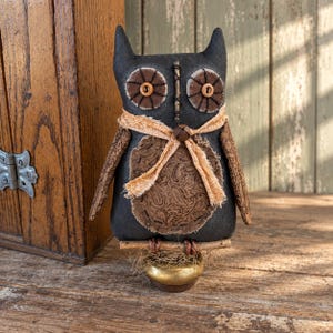 May include: A stuffed owl figurine made of black fabric with brown paisley patterned fabric on the chest and wings. The eyes are brown fabric circles with buttons in the center. A frayed tan ribbon is tied around the neck with a small metal bell.
