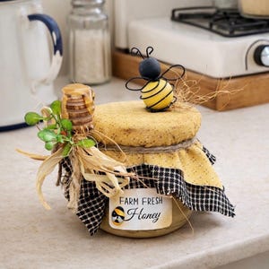 May include: A decorative honey jar with a yellow fabric lid, black and white checkered trim, and a small bee figurine on top. The jar has a label that reads "Farm Fresh Honey". A wooden honey dipper is placed on the side.