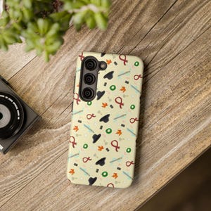 May include: A yellow phone case with a pattern of black hats, red ribbons, and green leaves. The case has a black camera lens cutout and a white border.