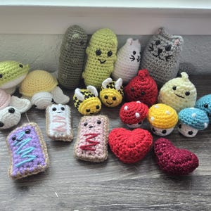 Op de afbeelding: Een verzameling gehaakte amigurumi-speelgoed, waaronder een verscheidenheid aan dieren, voedsel en andere objecten. De speelgoed is gemaakt in verschillende kleuren, waaronder groen, geel, rood, blauw en roze.