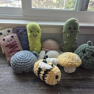 May include: A collection of crocheted plush toys, including a pickle, a mushroom, a bee, a snail, and a few other food-themed toys. The toys are all handmade and have cute faces.