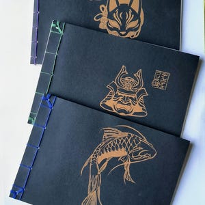 May include: Three black notebooks with exposed side stitching in purple, green, and blue. Each cover features a gold-colored line art illustration: a fox mask, a samurai helmet, and a koi fish, along with Japanese text in a square stamp.