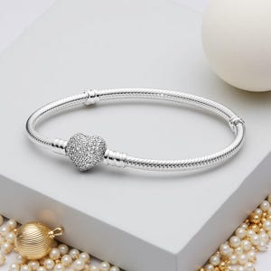 May include: A silver bracelet featuring a heart-shaped charm encrusted with small, sparkling stones. The bracelet has a textured, snake-chain design. A white sphere and gold-coloured jewellery are in the background.