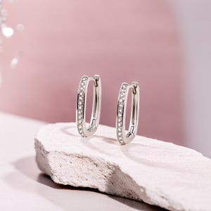 May include: Two silver hoop earrings, rectangular in shape, each set with small, clear gemstones. The earrings are presented on a light-coloured, textured surface, set against a soft pink backdrop. The earrings are a classic jewellery item.