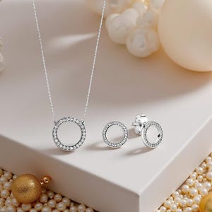 May include: A silver necklace and matching earrings set. The necklace has a circular pendant with a clear centre and a border of small, sparkling stones. The earrings have a similar design. The jewellery is displayed on a white surface.
