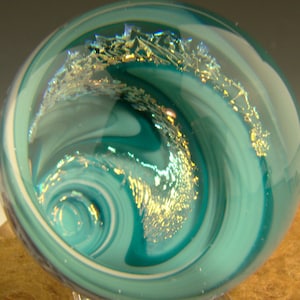 May include: A teal and white glass orb with gold flecks and a swirling pattern. The orb is sitting on a wooden surface.