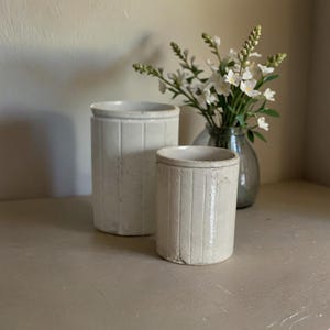 May include: Two white ceramic jars with a ribbed design. The larger jar is taller than the smaller jar. The jars are sitting on a light brown surface.