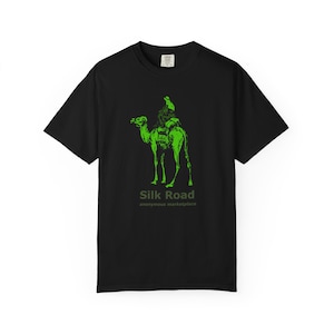 May include: A black short-sleeve t-shirt featuring a bright green graphic of a person riding a camel. Below the graphic, the text reads Silk Road in a bold font and anonymous marketplace in a smaller, thinner font underneath it.