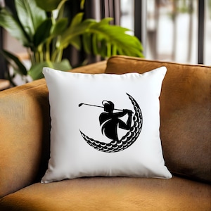 May include: White pillow with a black silhouette of a golfer swinging a club inside a crescent moon shape made of golf balls.