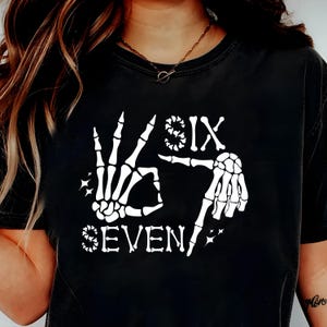 May include: Black t-shirt with a white skeleton hand graphic forming the number seven. The word "SIX" is above the hand, and the word "SEVEN" is below. Small starbursts accent the design.
