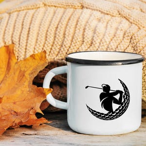 May include: White enamel mug with a black silhouette of a golfer hitting a golf ball in the shape of a crescent moon. The mug is sitting on a wooden surface with a brown knitted blanket and a fall leaf in the background.