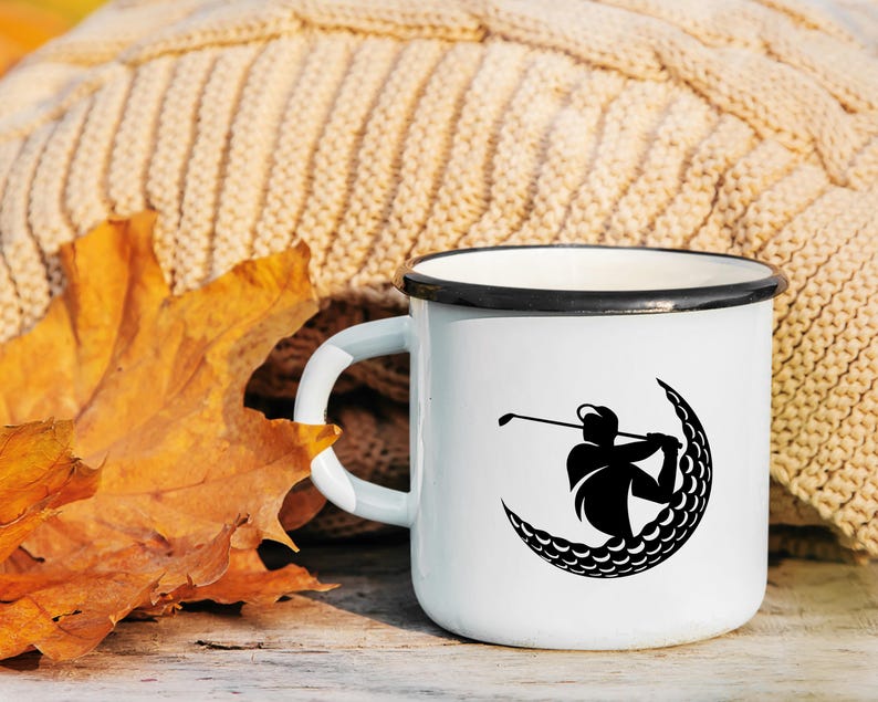 May include: White enamel mug with a black silhouette of a golfer hitting a golf ball in the shape of a crescent moon. The mug is sitting on a wooden surface with a brown knitted blanket and a fall leaf in the background.